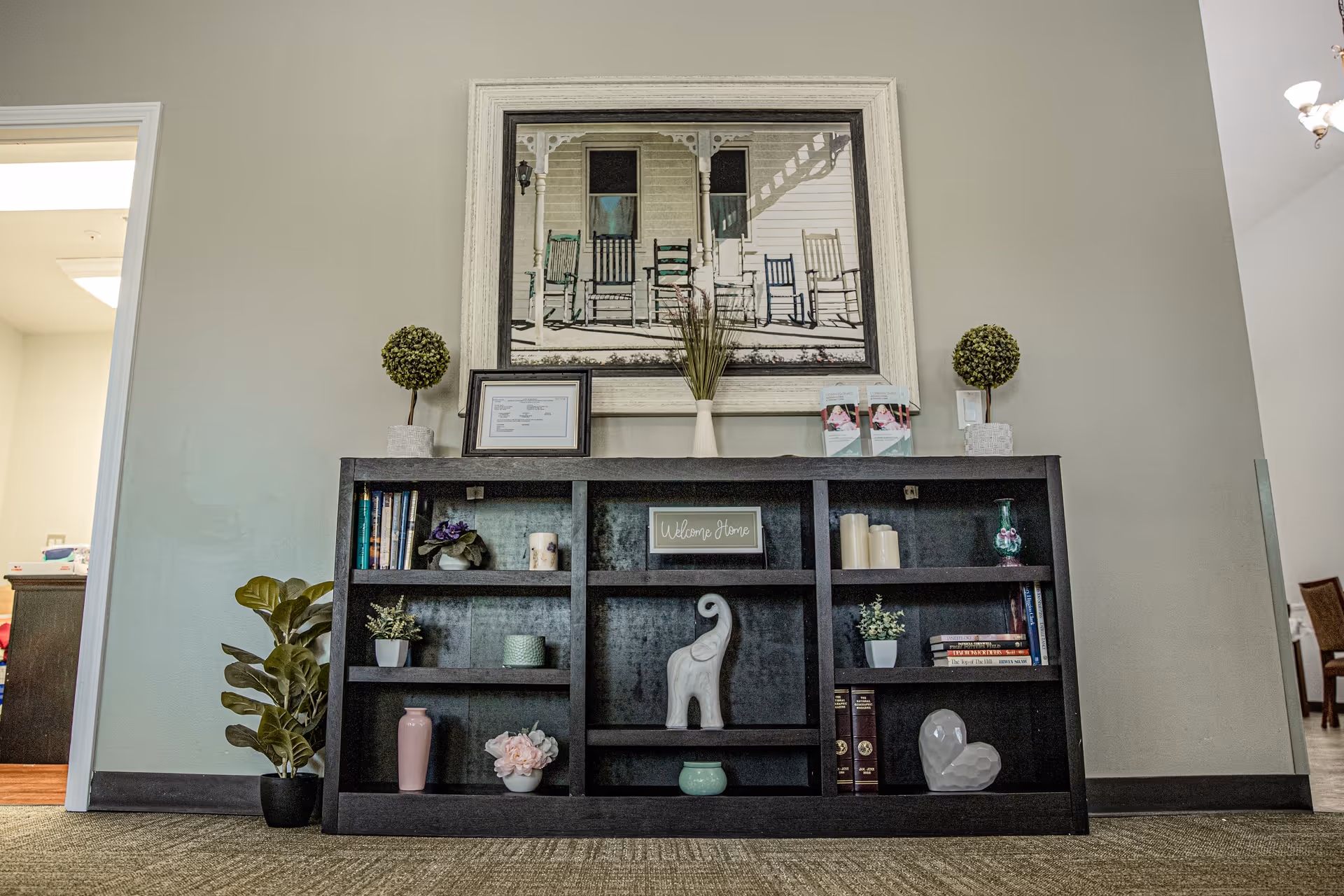 A black bookshelf against a beige wall with various decorative items including small plants, books, candles, and figurines. Above the bookshelf is a large framed picture of a porch with rocking chairs. There are two small potted topiary plants on top of the bookshelf, along with a framed certificate and some brochures. The room has carpeted flooring and doorways leading to other rooms on either side.