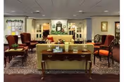 A cozy living room area in a senior living facility with a yellow sofa, wooden table with decorative items, four upholstered armchairs, a patterned rug, and festive holiday decorations including garlands and a wreath on the wall.