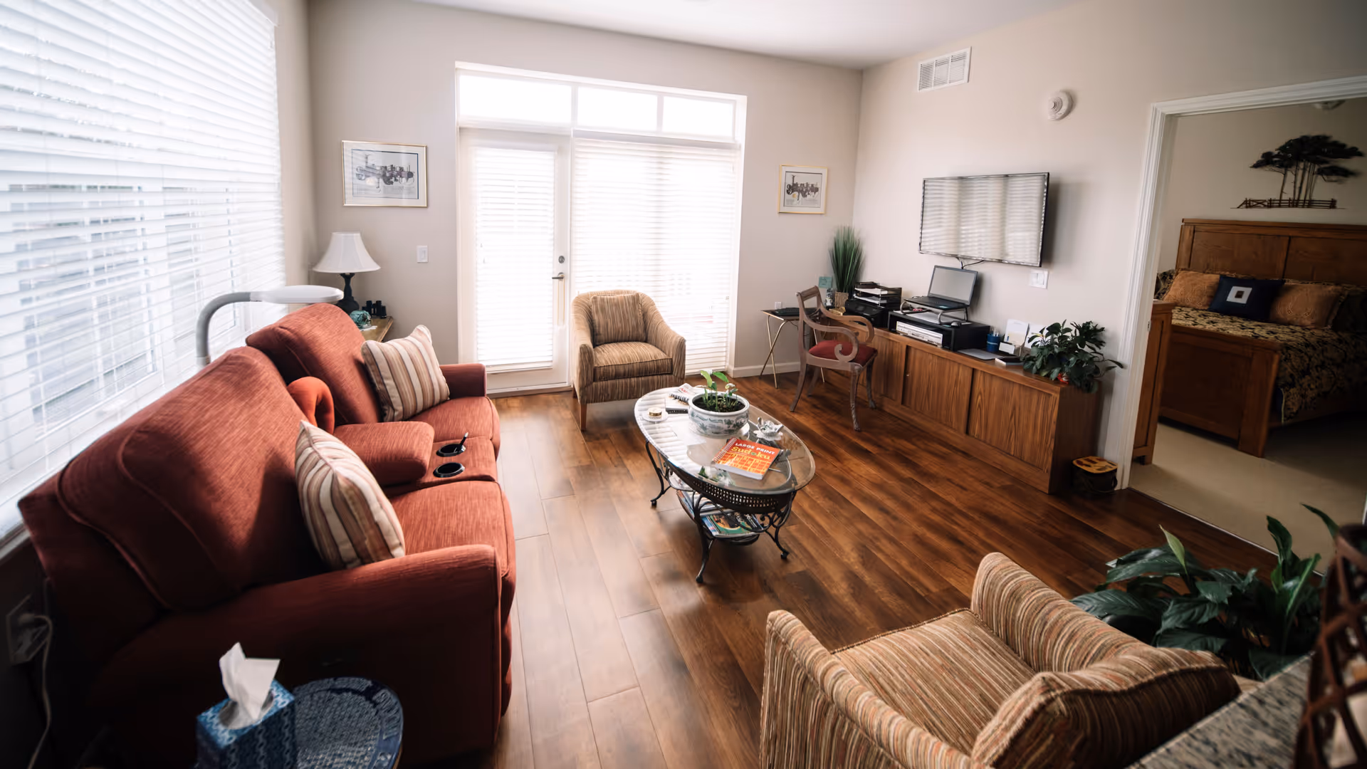 Bright living room with a red sofa, armchairs, a glass coffee table, wall-mounted TV, and an open doorway to a bedroom.
