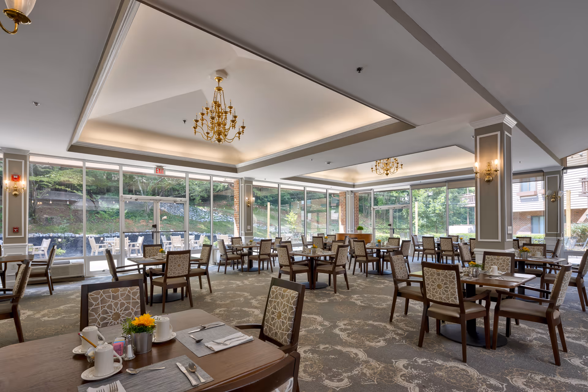A spacious dining room with multiple wooden tables and chairs arranged neatly. The room features large floor-to-ceiling windows allowing natural light to fill the space and offering a view of an outdoor patio area with additional seating. The ceiling has recessed lighting and elegant gold chandeliers. The carpet has a decorative pattern, and the tables are set with placemats, utensils, cups, and small flower arrangements.
