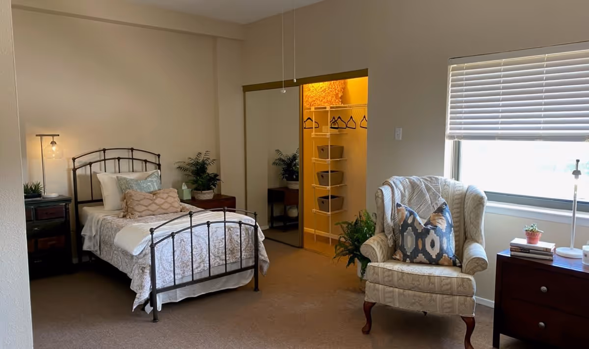 A cozy bedroom with a single metal bed frame, neatly made with pillows and a quilt. There is a nightstand with a lamp and a plant next to the bed. A mirrored closet door is open, revealing shelves with baskets and hangers. An upholstered armchair with a decorative pillow sits near a window with blinds, next to a small wooden dresser with books and a small plant on top.