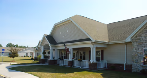 Exterior view of a single-story senior living facility building with beige siding, a covered porch with white railings, an American flag, and a well-maintained lawn under a clear blue sky.