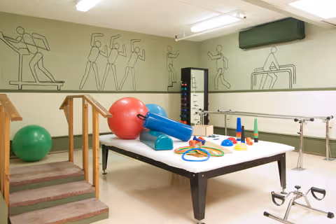 A physical therapy or rehabilitation room with exercise equipment including a large table with therapy balls, rings, cones, and other small exercise tools. There are parallel bars along the wall for walking exercises, a set of stairs with handrails, and a green exercise ball on the floor. The walls are decorated with line art illustrations of people exercising.