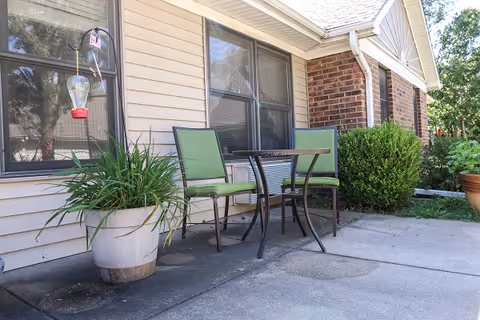 Outdoor patio area with two green cushioned chairs and a small metal table on a concrete surface next to a beige and brick building. There is a large potted plant with a hummingbird feeder hanging above it and some bushes along the building.