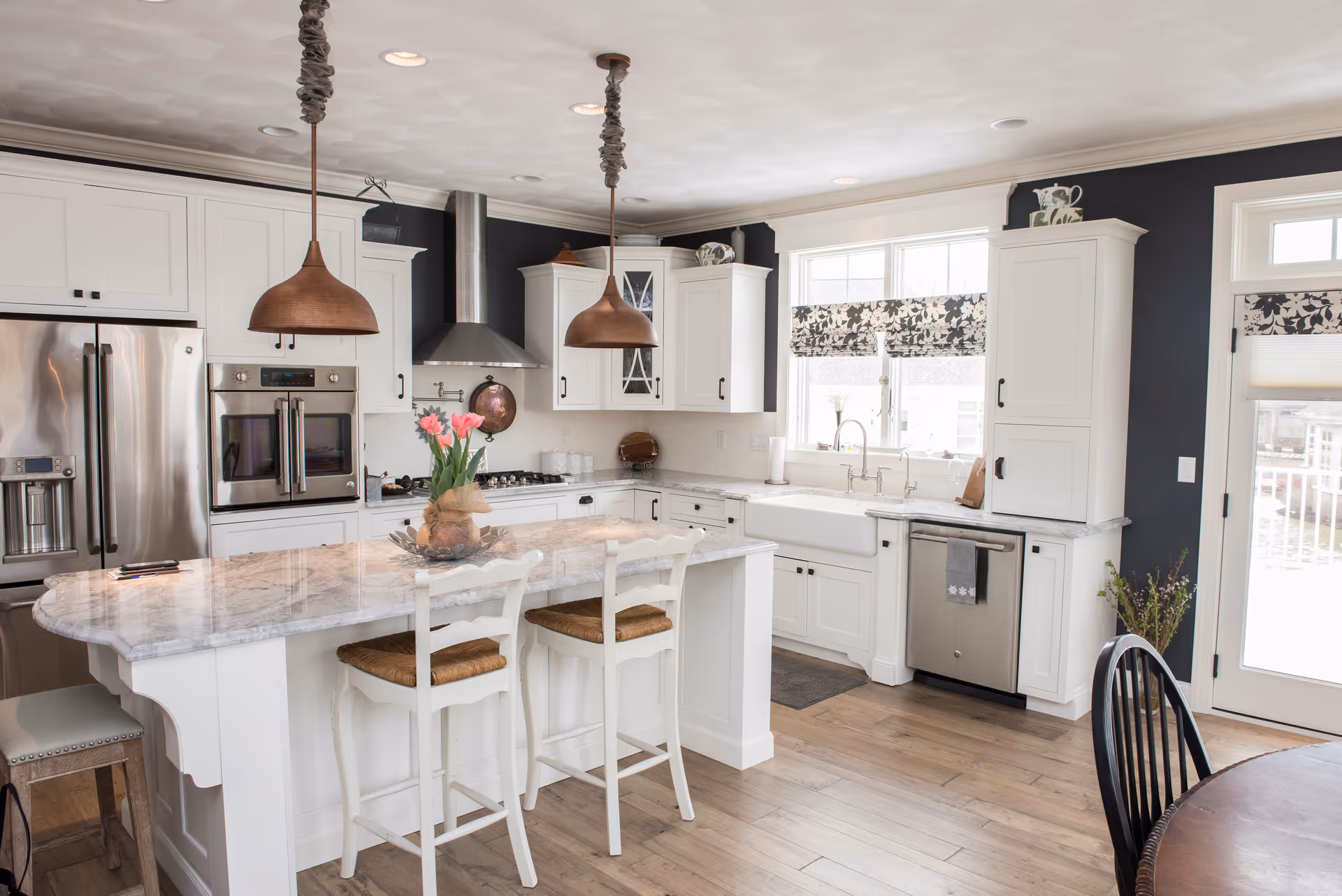 Bright and modern kitchen with white cabinetry, stainless steel appliances including a refrigerator, double oven, and dishwasher. A large marble island with two white wooden chairs with woven seats is in the center. Two copper pendant lights hang above the island. There are floral-patterned window shades on the windows, and a door with glass panels leads outside. The floor is light wood, and a vase with pink flowers is on the island.