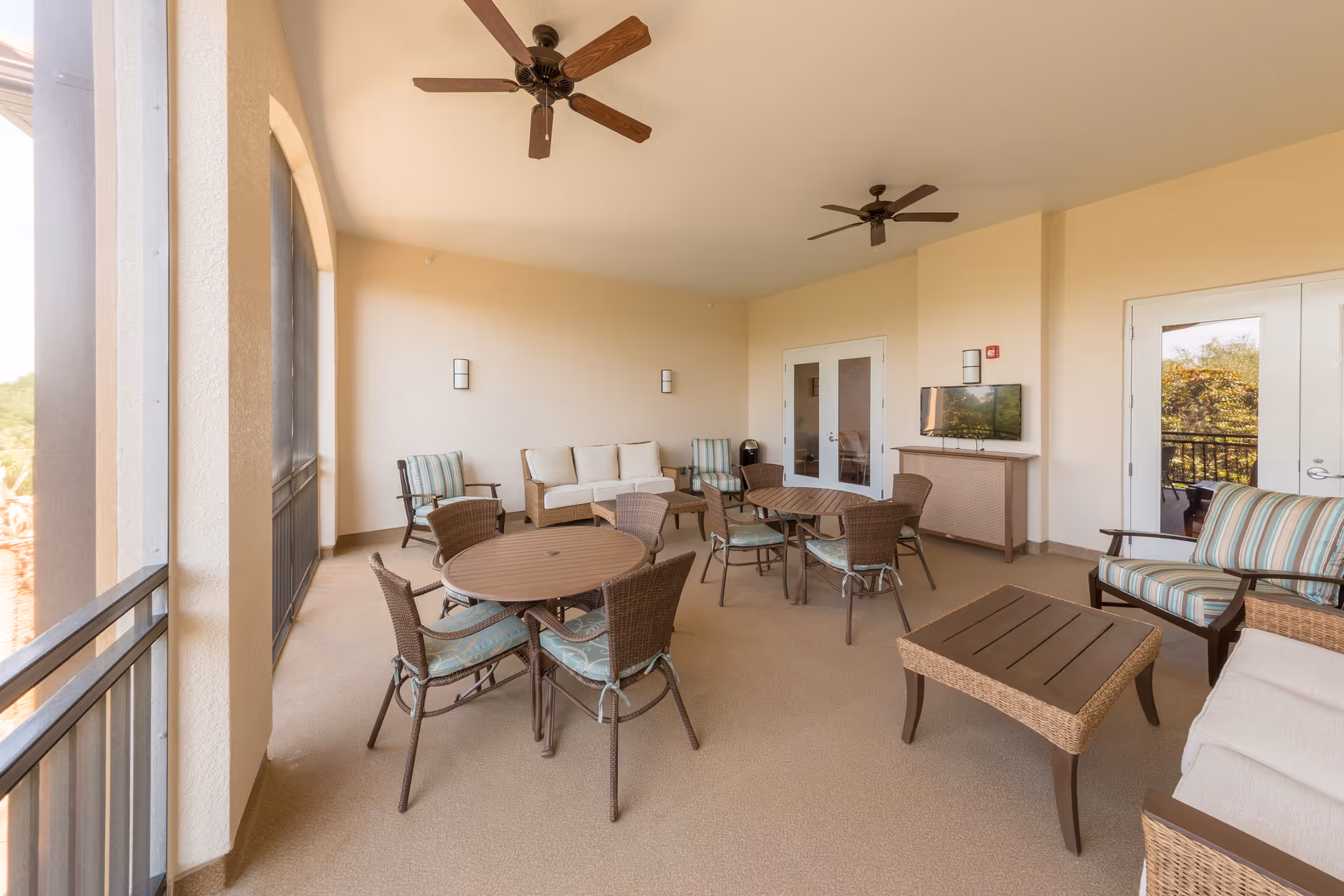 A spacious covered patio area with multiple round tables and wicker chairs with cushions. There are ceiling fans, a wall-mounted TV, and comfortable seating including a sofa and armchairs with striped cushions. The area is enclosed with large screened windows and has double glass doors leading inside.