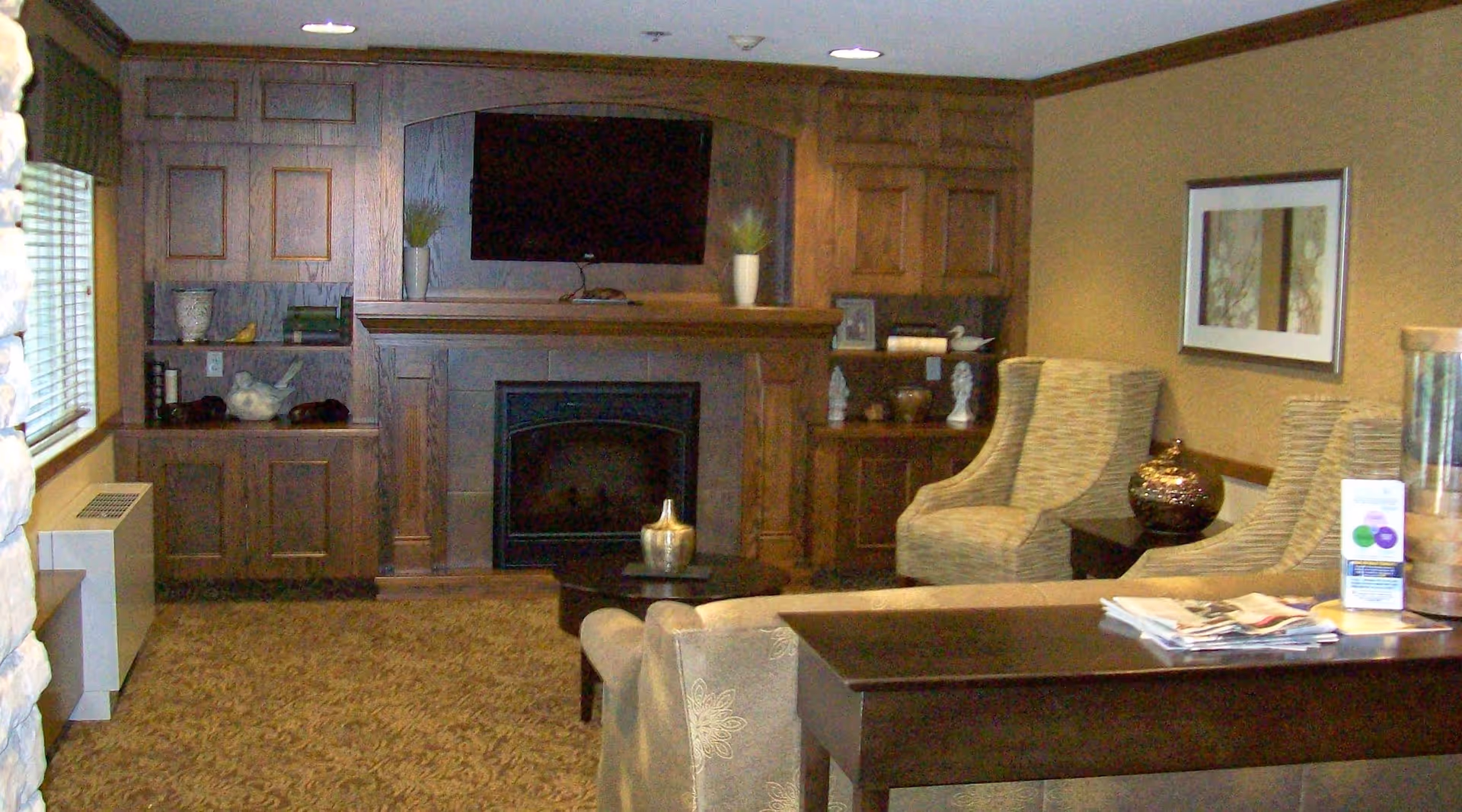 A cozy living room area with a fireplace and a mounted flat-screen TV above it. The room features built-in wooden cabinets on either side of the fireplace, two upholstered armchairs, a dark wooden table with magazines on it, and a framed picture on the wall. The carpet has a patterned design and there is a window with blinds on the left side.