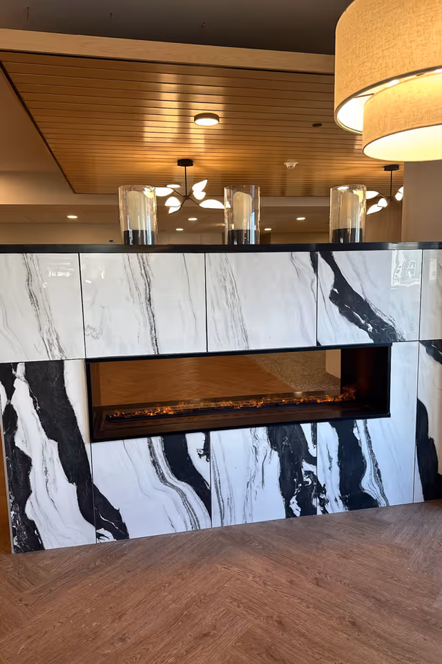 Modern marble-tiled double-sided fireplace in a lobby area with wood ceiling panels and pendant lights.