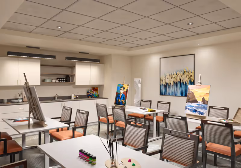 A well-lit art room with several tables and chairs arranged for painting activities. There are easels with colorful paintings, paintbrushes, and paint bottles on the tables. The room has white cabinets and shelves along one wall, and a large abstract painting hangs on another wall.