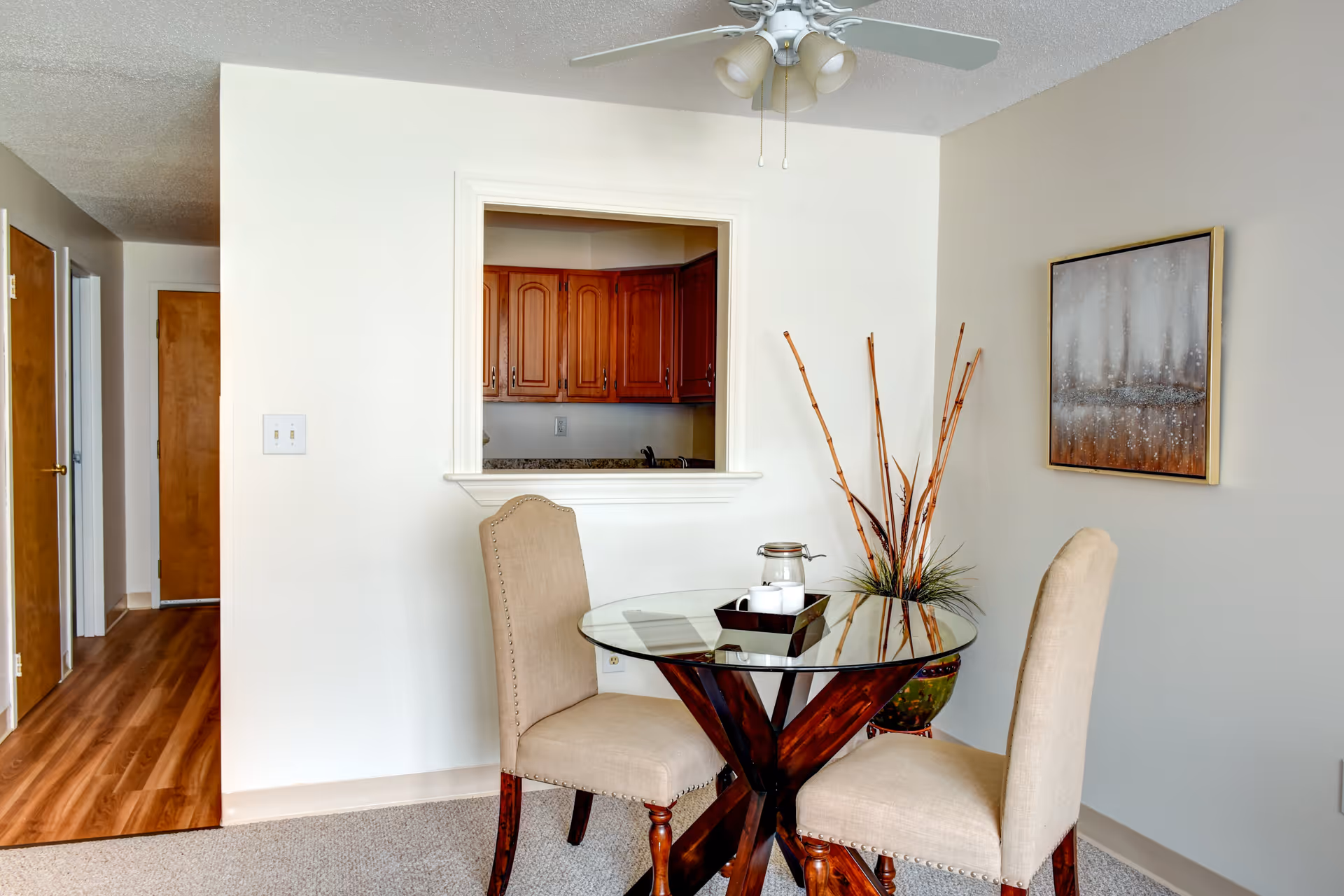 A small dining area with a round glass table and two beige upholstered chairs. On the table is a decorative tray with a candle and a small jar. Behind the table is a wall with a pass-through window showing a kitchen with wooden cabinets. A tall vase with decorative sticks and a framed abstract painting are on the right side. The floor is carpeted and there is a ceiling fan with lights above.