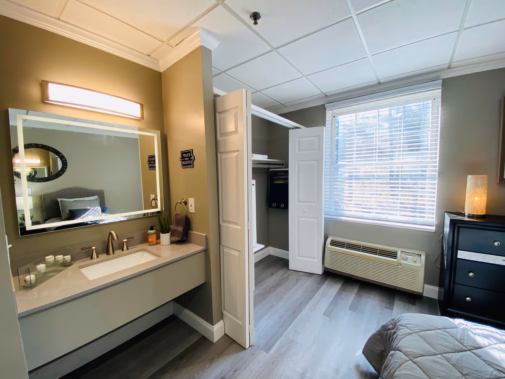 A senior living facility bedroom with a bed partially visible on the right, a black dresser with a lit lamp on top, a window with white blinds, an air conditioning unit below the window, and an open closet with white bi-fold doors. On the left side, there is a bathroom sink with a large illuminated mirror above it, a soap dispenser, a small plant, and a towel hanging on a ring.