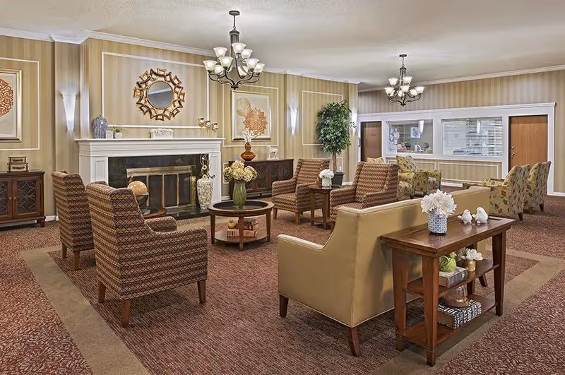 A cozy living room area in a senior living facility with multiple armchairs and sofas arranged around a central coffee table. The room features a fireplace with decorative items on the mantel, framed artwork on the walls, a chandelier, and a side table with flowers and decorative objects. The walls have a striped wallpaper and the carpet is a warm brown color.
