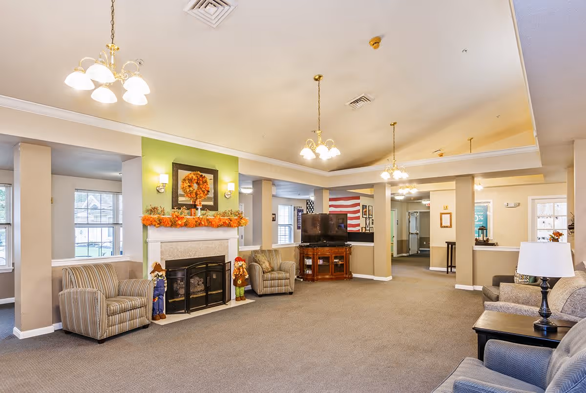 A spacious senior living facility common area with beige walls and carpeted floor. The room features a fireplace decorated with autumn-themed garland and a wreath above it. Two striped armchairs flank the fireplace, and a wooden TV stand with a flat-screen TV is positioned against a wall. Several ceiling light fixtures hang from the ceiling, and additional seating with armchairs and a side table with a lamp are visible. Large windows allow natural light into the room.