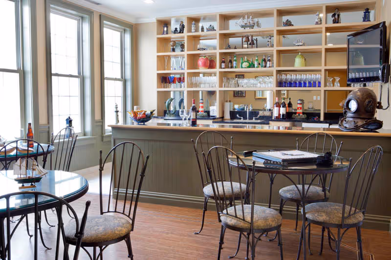 A cozy interior room with a bar counter and shelves filled with decorative items and glassware. There are several round tables with metal chairs featuring cushioned seats. Large windows allow natural light to brighten the space, and a vintage diving helmet is displayed on the bar counter.