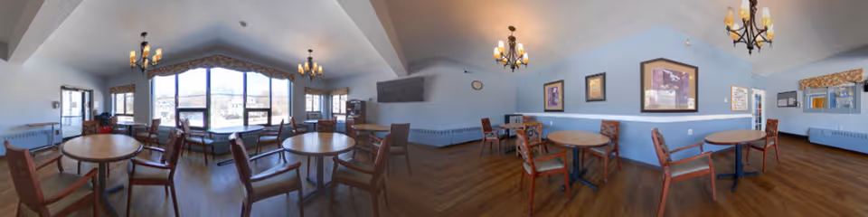 A spacious dining room with multiple round wooden tables and chairs arranged neatly. The room has large windows allowing natural light to fill the space, light blue walls with framed artwork, and chandeliers hanging from the ceiling. The floor is wooden with a herringbone pattern.