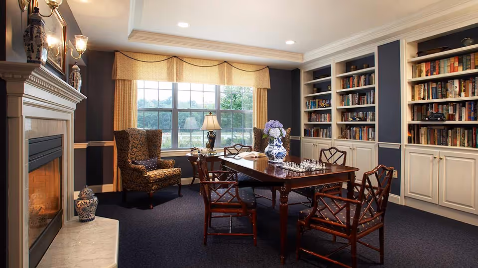A cozy library room with a large window covered by beige curtains, a wooden table with a chess set and a blue and white vase with flowers, surrounded by four wooden chairs with patterned cushions. There is a patterned armchair near the window, a table lamp, built-in bookshelves filled with books, and a fireplace with decorative vases on the mantel.