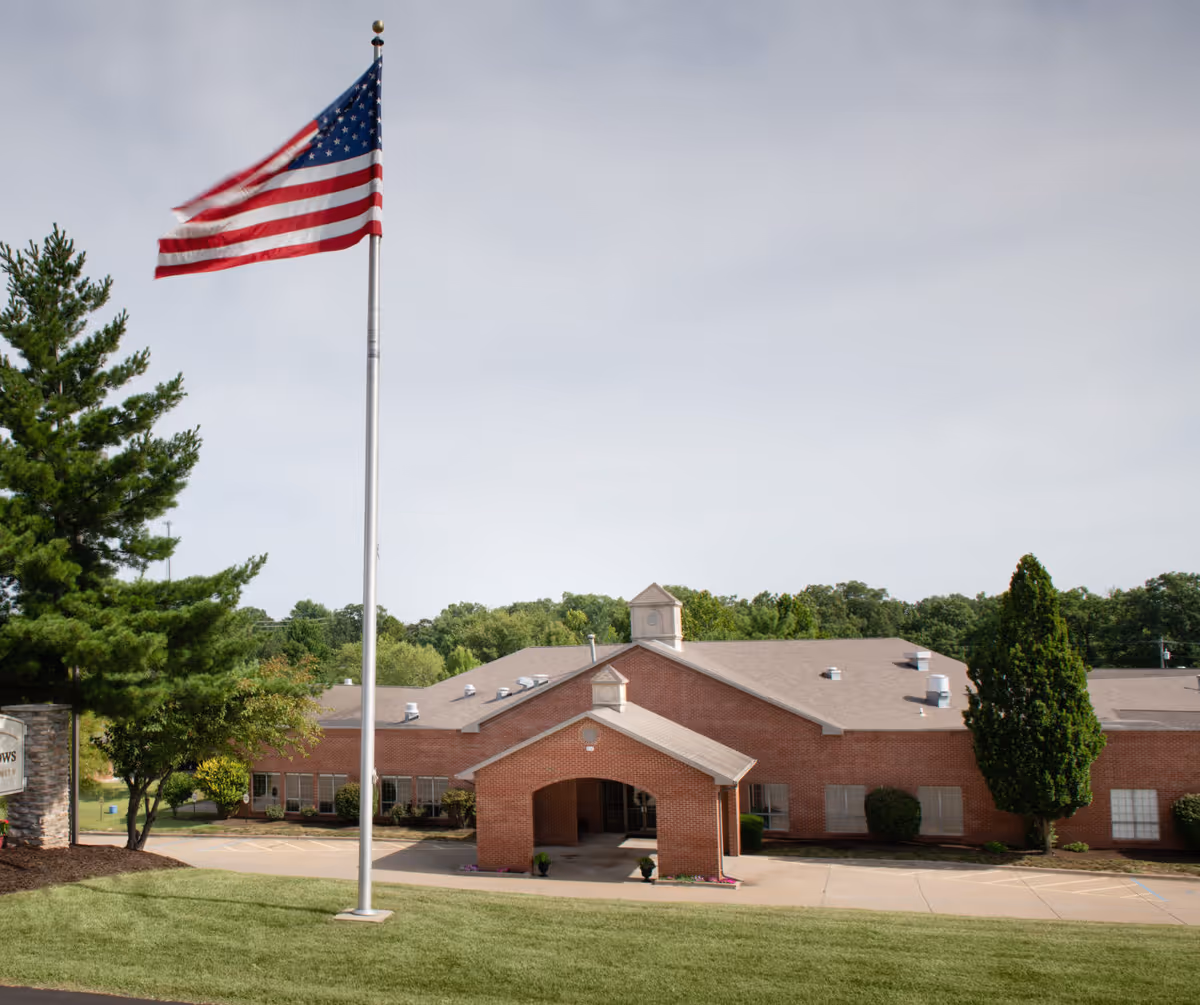 Exterior view of a single-story brick building with a covered entrance, surrounded by green trees and grass. An American flag on a tall flagpole is prominently displayed in the foreground against a partly cloudy sky.