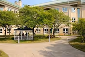 Outdoor courtyard area of a senior living facility with a paved walkway, green trees, and a white gazebo. The building with multiple windows is visible in the background under a clear sky.