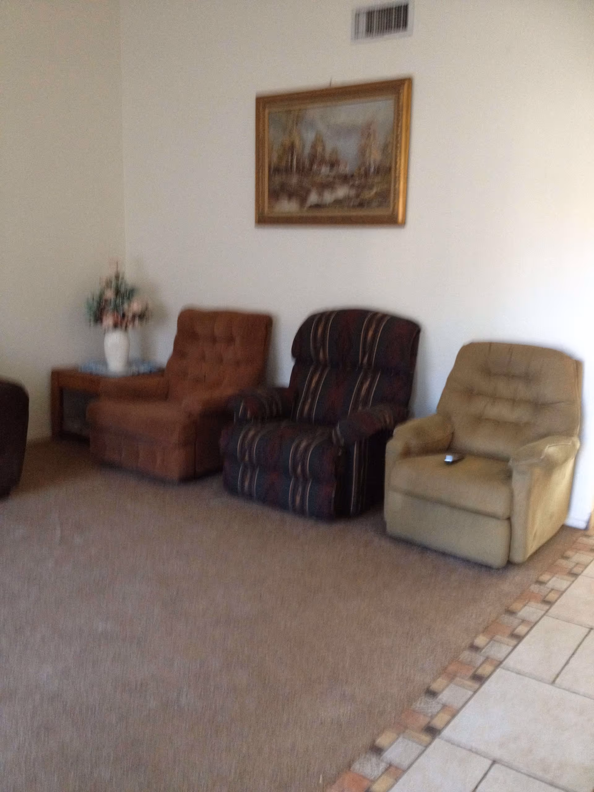 A living room area with three upholstered armchairs in different colors arranged against a white wall. Above the chairs hangs a framed painting depicting a landscape scene. To the left of the chairs is a small wooden side table with a white vase holding a bouquet of flowers. The floor is carpeted with a tiled border visible on the right side.