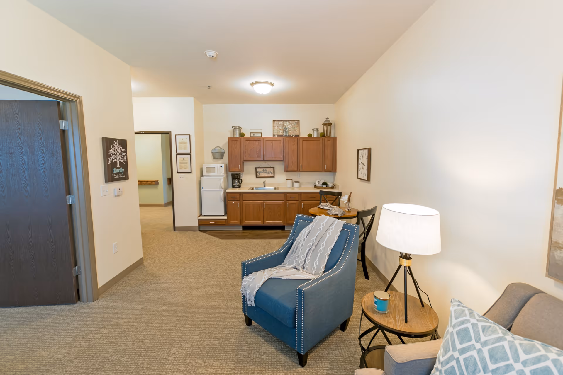 Open living area featuring a blue armchair, side table and lamp with a small kitchenette and wooden cabinets in the background.