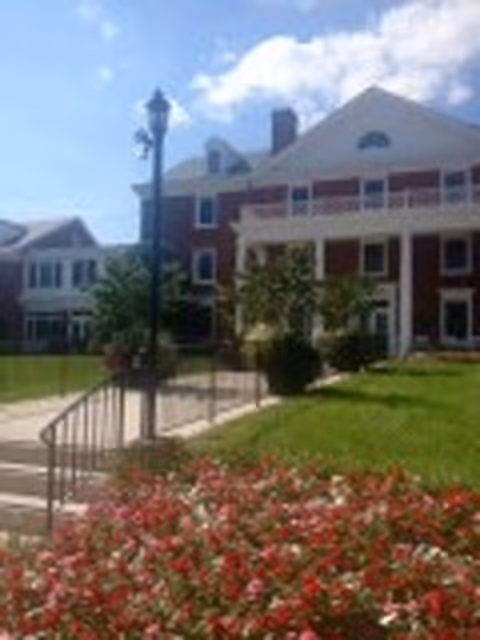 Exterior view of a large brick building with white columns and a porch, surrounded by green grass and flower beds with red and white flowers under a partly cloudy blue sky.