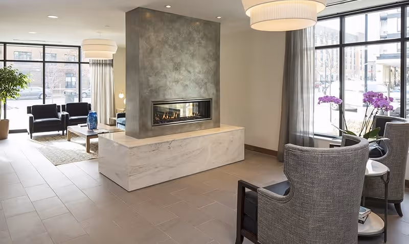 A modern living room area with a central fireplace featuring a concrete and marble surround. The room has large windows letting in natural light, with gray armchairs and a small round table with purple orchids near the window. In the background, there are black armchairs and a wooden coffee table on a rug.