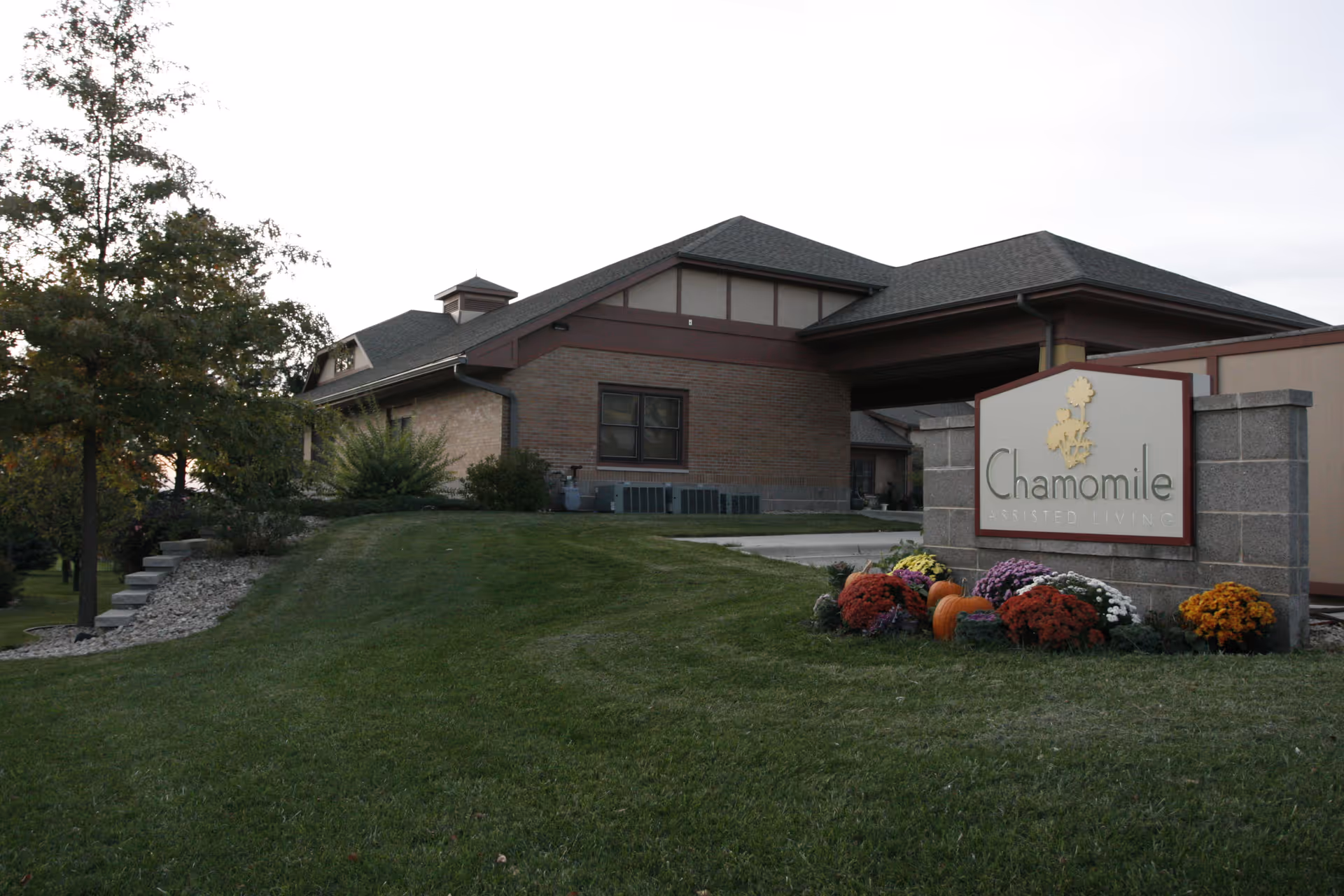 Exterior view of Chamomile Assisted Living Ltd building with a manicured lawn, trees, and a sign displaying the facility's name surrounded by colorful flowers and pumpkins.