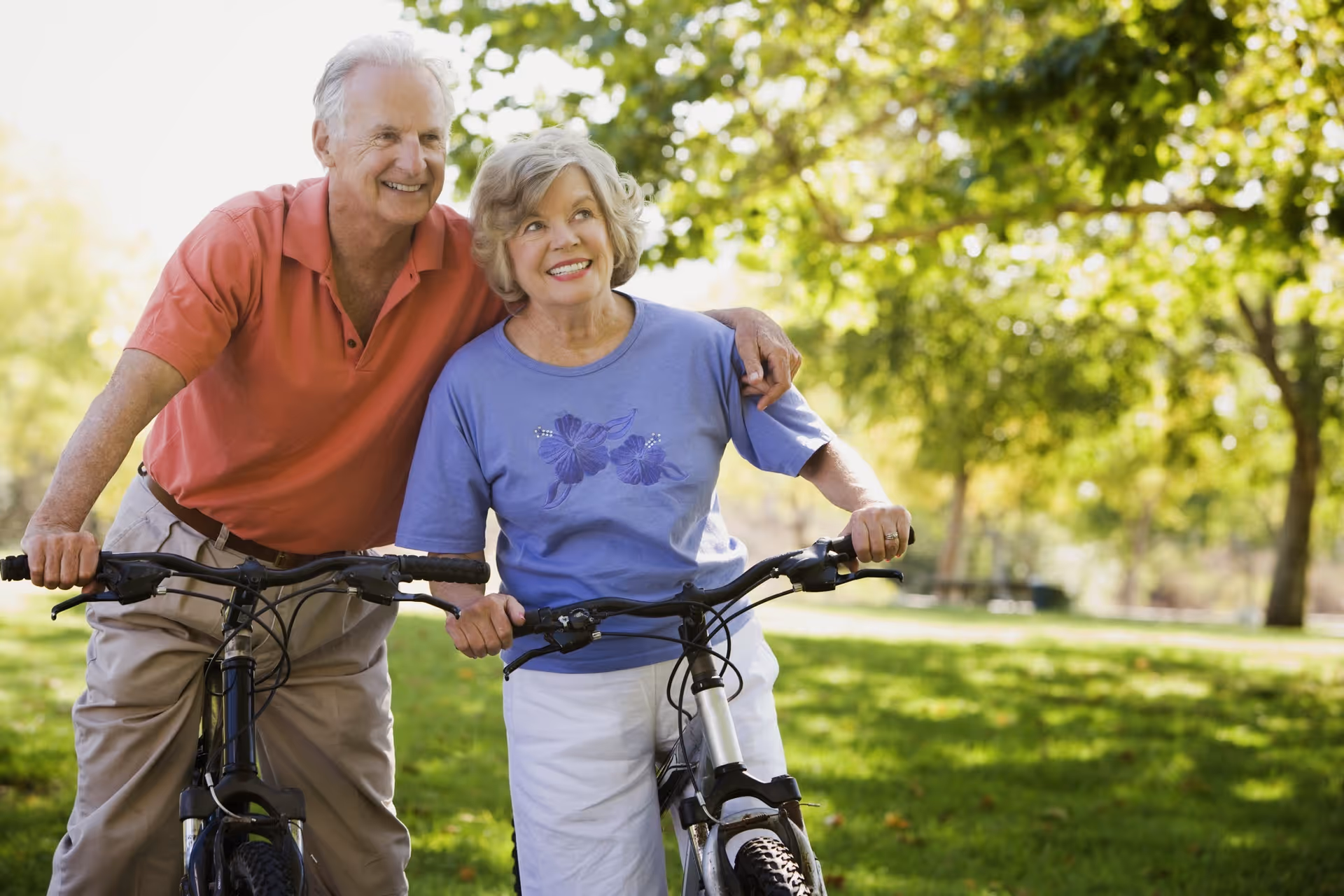 An elderly couple outdoors in a park, each holding a bicycle. The man is wearing a red polo shirt and beige pants, smiling and looking at the woman. The woman is wearing a blue shirt with floral designs and white pants, smiling and looking slightly to the side. They are surrounded by green grass and trees with sunlight filtering through.