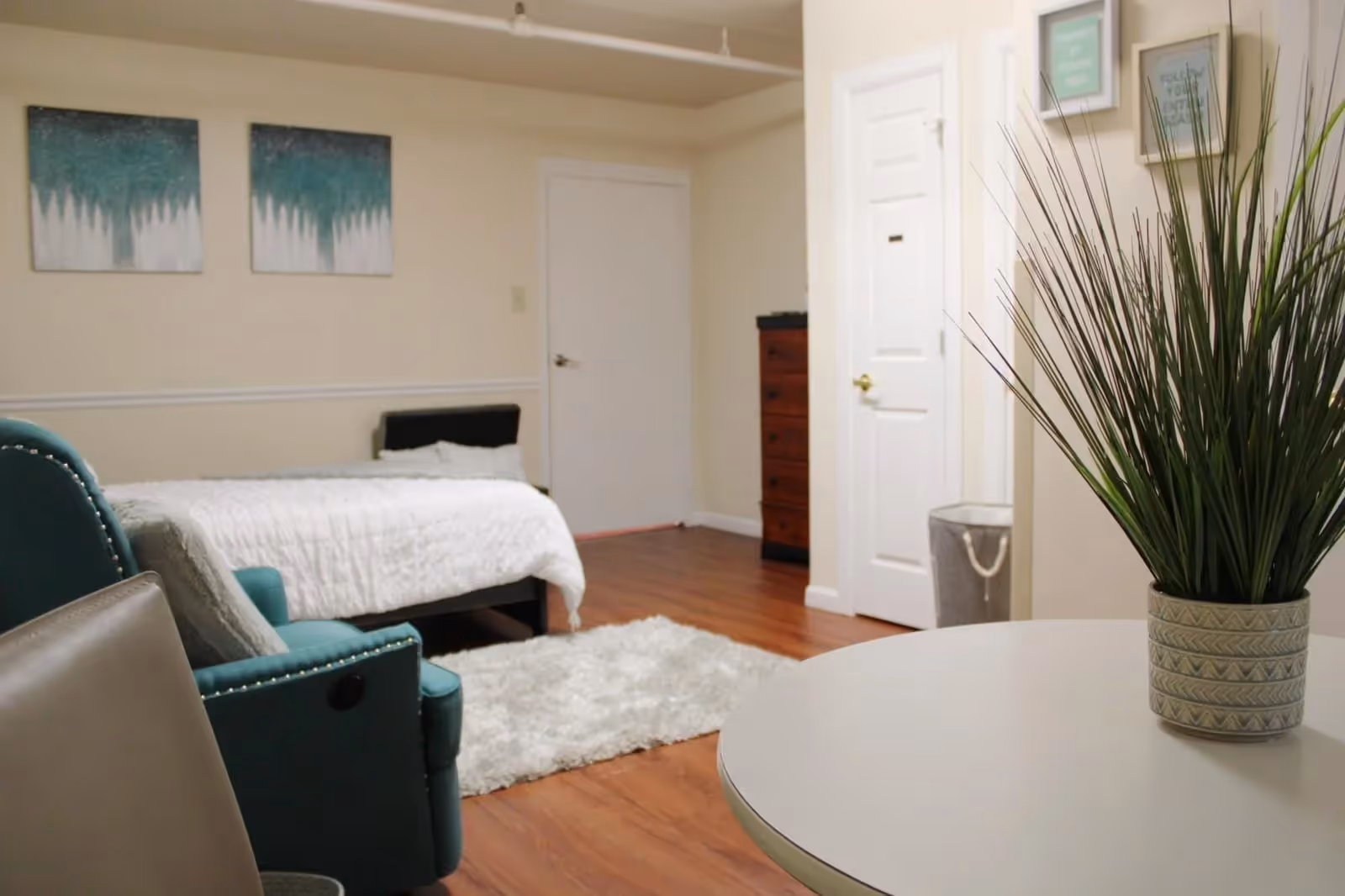 A cozy bedroom with a single bed covered in a white quilt, a teal armchair, a white fluffy rug on wooden flooring, a small round table with a potted plant, and two abstract paintings on the wall.
