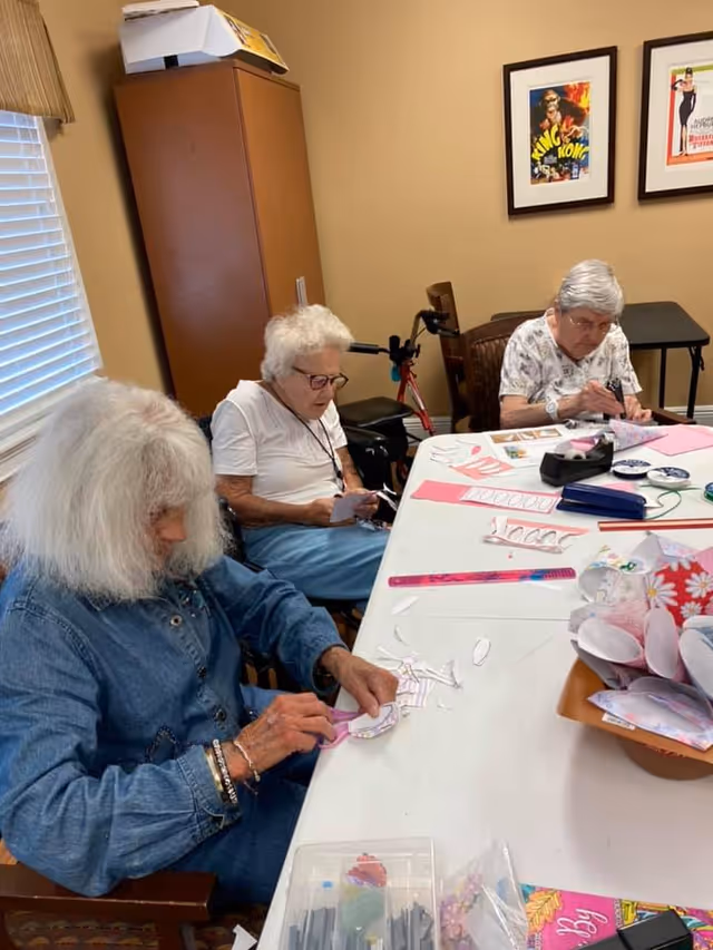 Three elderly women sitting around a table engaged in arts and crafts activities, cutting and working with paper in a well-lit room with beige walls and framed artwork on the wall.