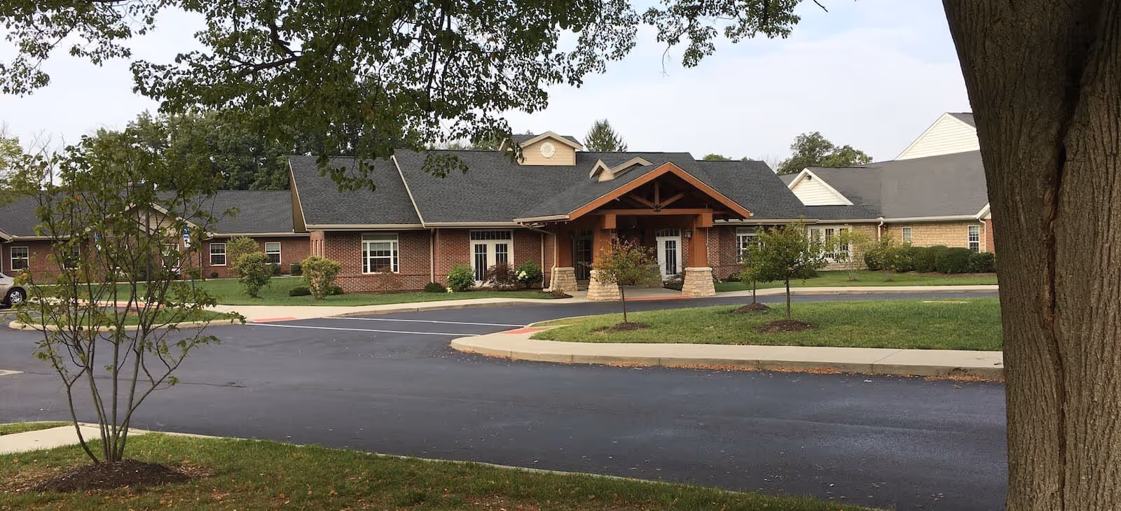 Front exterior view of a single-story brick and siding building with a covered entrance supported by stone pillars, surrounded by a paved driveway, small trees, and green lawn under a partly cloudy sky.