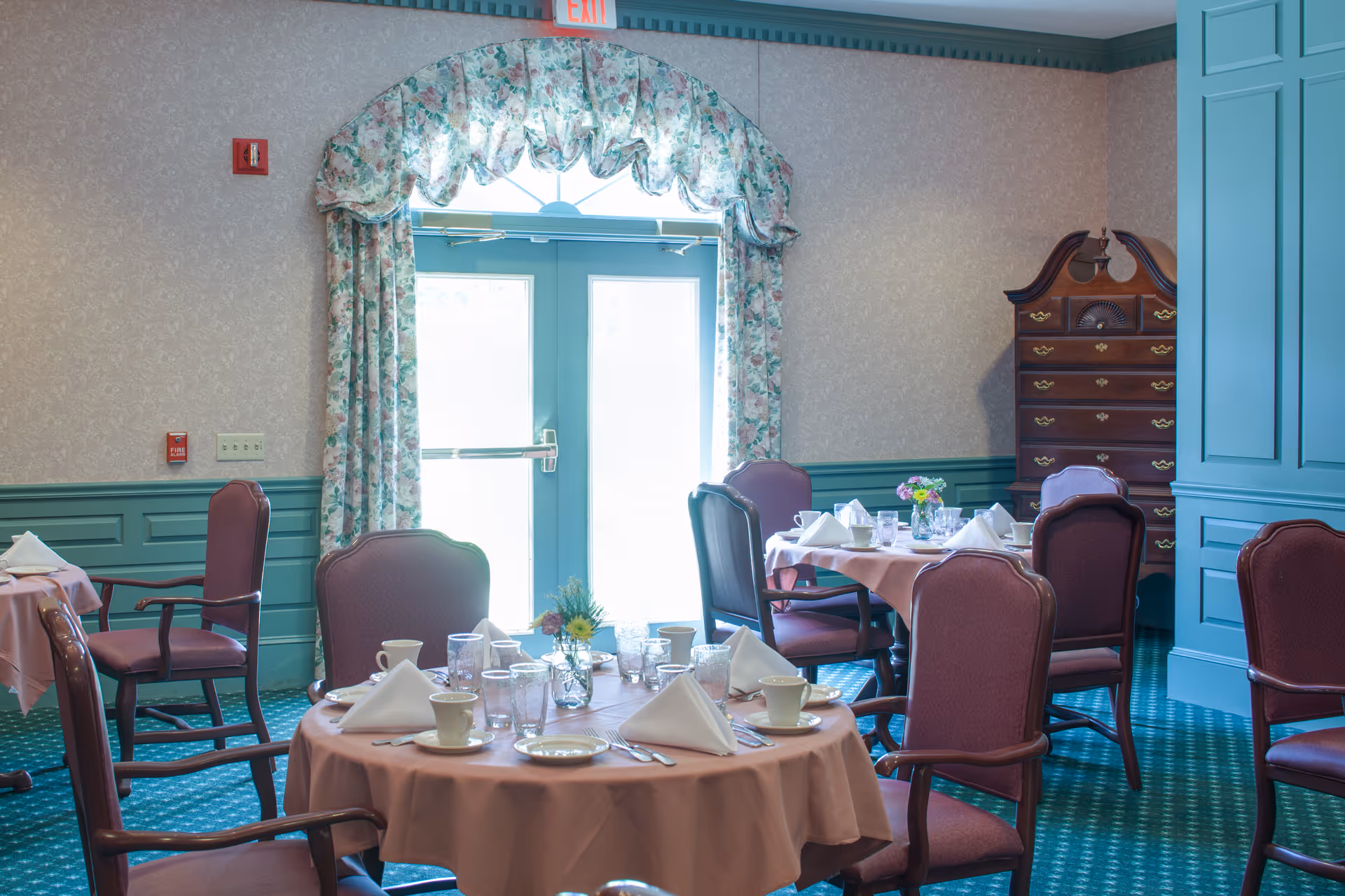 A dining room with round tables covered in pink tablecloths, set with white napkins, cups, glasses, and silverware. The room features teal wainscoting and trim, floral curtains framing a glass door with an exit sign above, and a tall wooden chest of drawers in the corner.