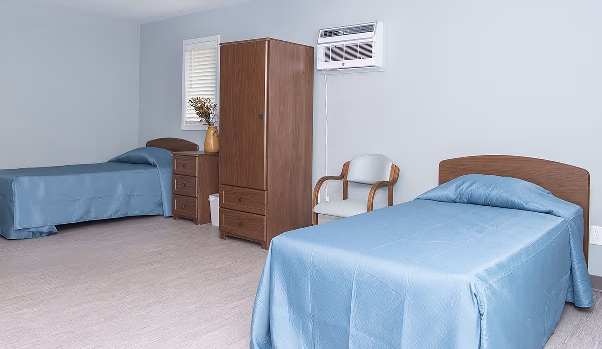 A simple, clean bedroom with two single beds covered in light blue bedspreads. Between the beds is a wooden dresser with a vase of flowers on top, and next to it is a wooden wardrobe. A white chair with wooden armrests is placed near the bed on the right. The walls are painted light gray, and there is a window with white blinds and a wall-mounted air conditioning unit above the chair.