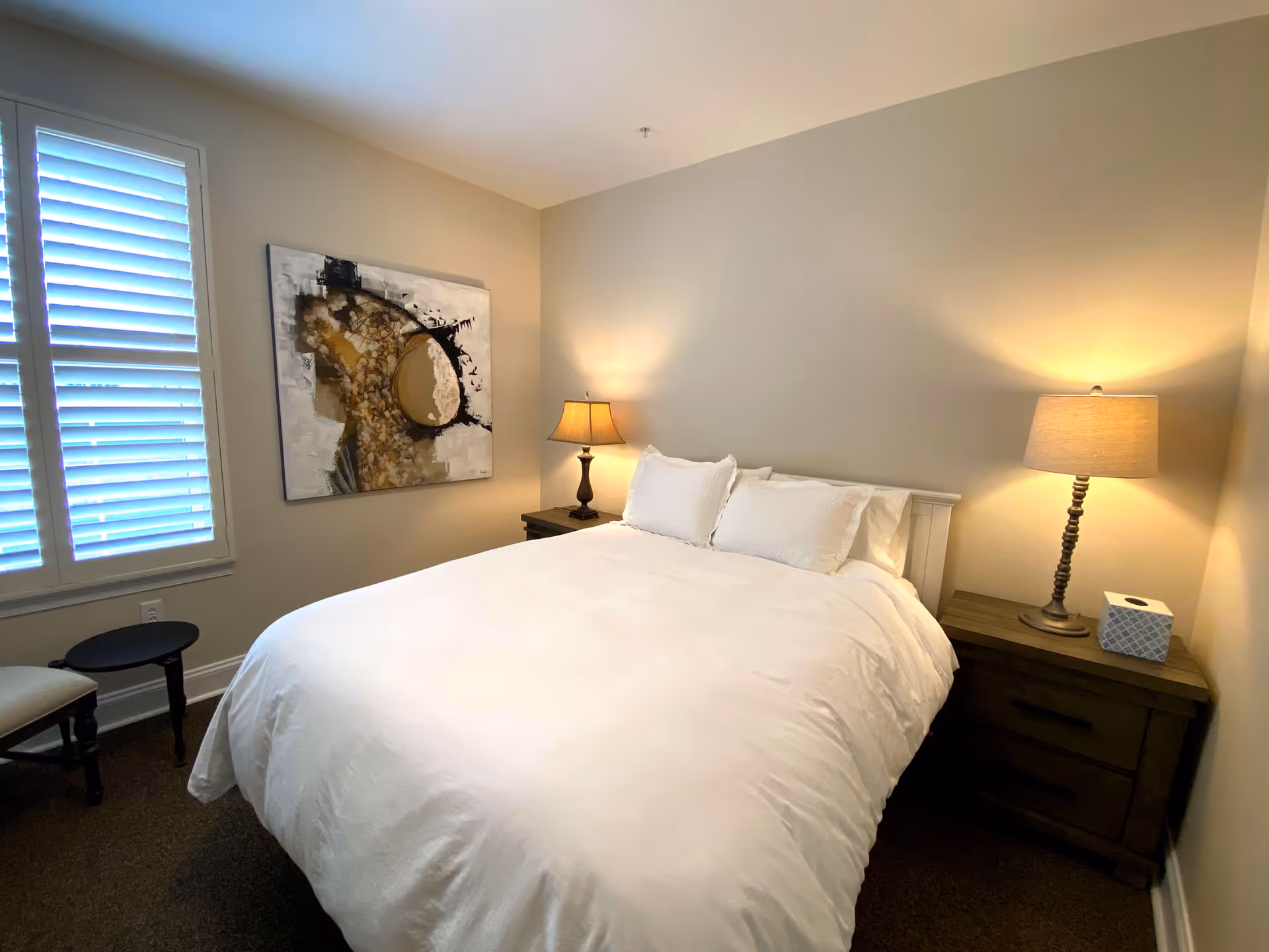 A bedroom with a neatly made bed featuring white bedding and three pillows. There are two wooden nightstands on either side of the bed, each with a lamp. A box of tissues is on the right nightstand. A window with white shutters is on the left wall, and an abstract painting hangs on the wall above the left nightstand.