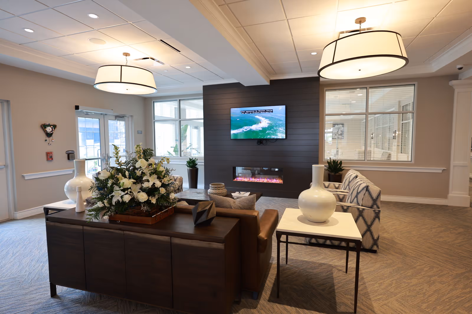 A modern senior living facility common area with comfortable seating including a leather sofa and patterned armchairs. A dark wood console table with a large floral arrangement and decorative vases is in the foreground. The room features a wall-mounted flat screen TV above a sleek electric fireplace. Large windows and glass doors allow natural light to fill the space, and two large ceiling light fixtures provide additional illumination.