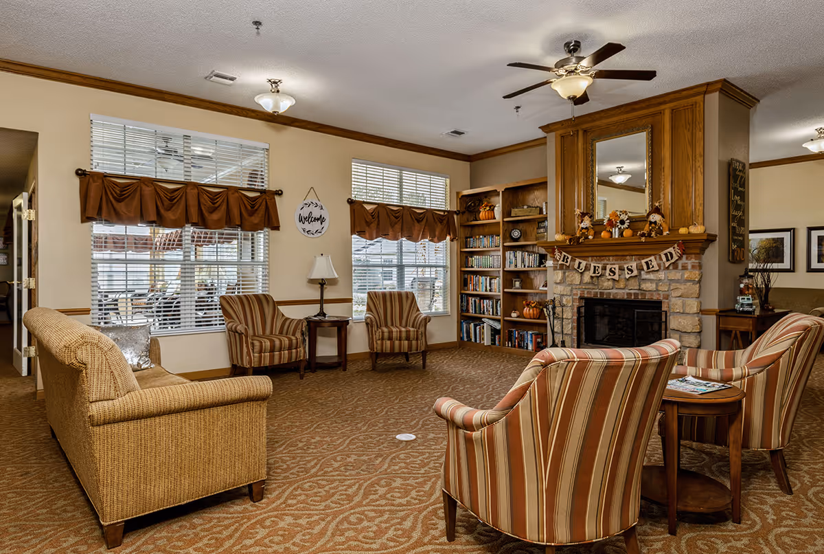 A cozy living room area in a senior living facility with a stone fireplace decorated with autumn-themed items and a 'BLESSED' banner. The room features a beige patterned carpet, a beige sofa, four striped armchairs arranged around small wooden tables, two large windows with brown valances, a bookshelf filled with books, and ceiling fans with lights.