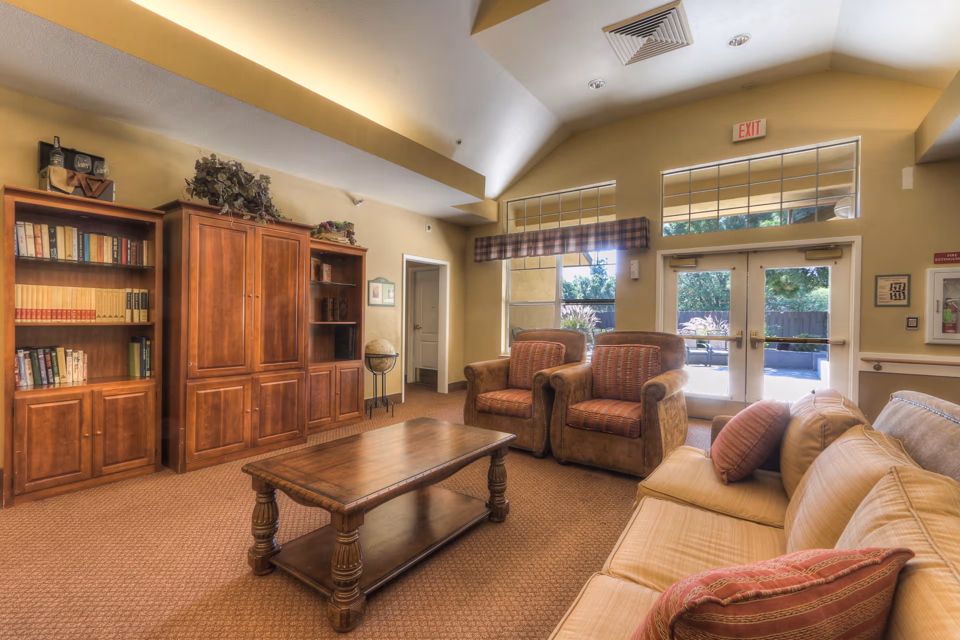 A cozy living room area with a beige sofa adorned with red and beige striped pillows, two brown armchairs with patterned cushions, and a wooden coffee table in the center. Behind the seating area, there are large windows and glass doors letting in natural light, with a view of outdoor greenery. On the left side, there are wooden bookshelves filled with books and decorative items, and a globe on a stand. The walls are painted a warm beige color, and the ceiling is vaulted with recessed lighting.