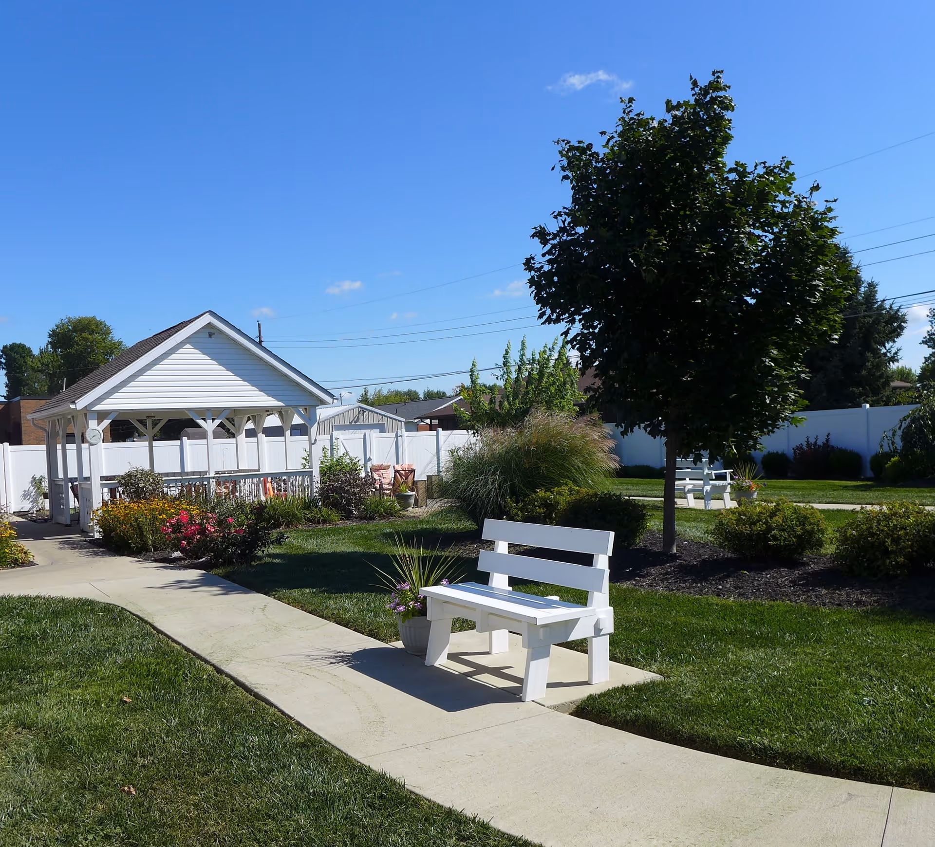 Sunny landscaped courtyard with a white gazebo, benches, walking path, and trees.