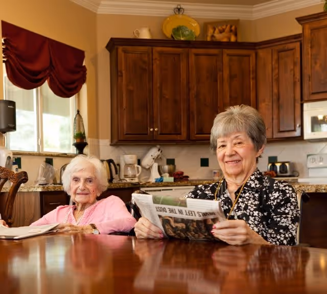 Two elderly women sitting at a wooden table in a kitchen. One woman is wearing a pink sweater and the other is wearing a black and white patterned blouse, holding a newspaper. The kitchen has wooden cabinets, a coffee maker, and a window with a red valance.