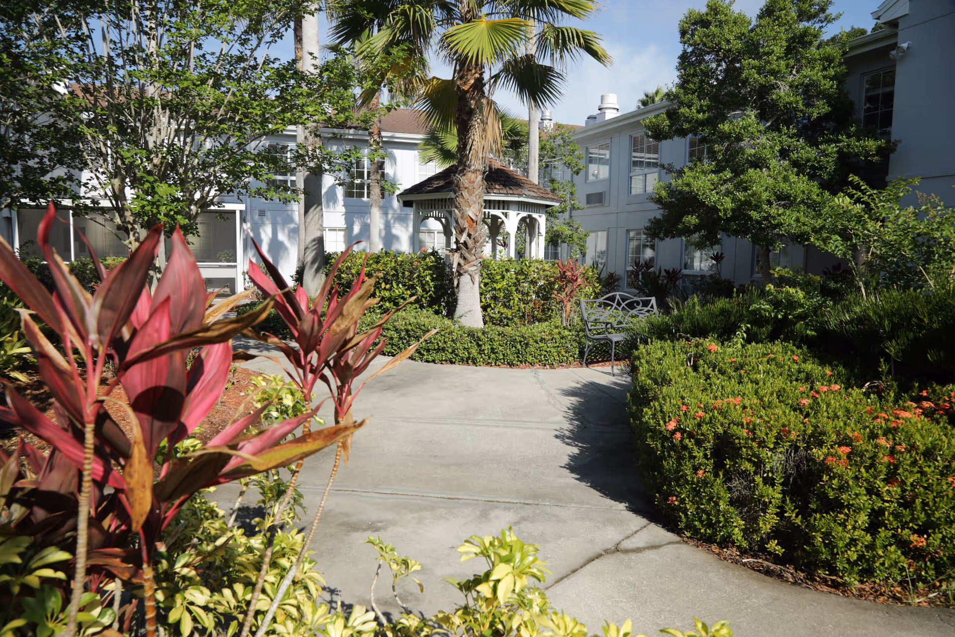 A sunny outdoor garden area with a concrete pathway winding through lush green bushes, colorful plants, and trees. There is a white gazebo in the background and a metal bench along the path. The building of the facility is visible behind the garden.