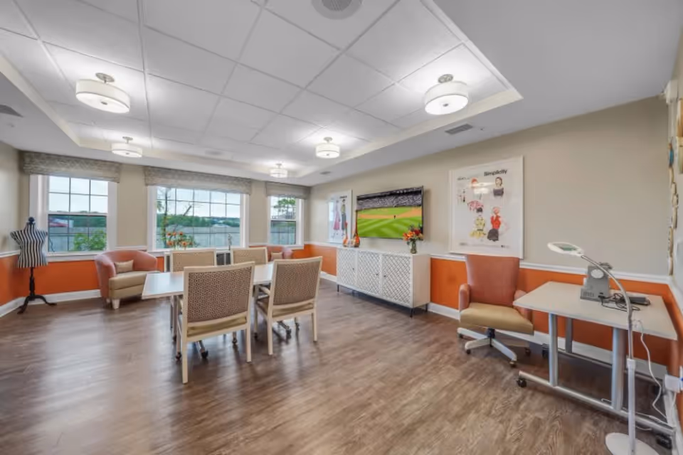 A bright and spacious common area with large windows letting in natural light. The room features a table with four chairs in the center, two armchairs near the windows, a TV mounted on the wall displaying a baseball game, and a small desk with a lamp and chair. The walls are painted beige with an orange accent below the chair rail, and there are framed artworks on the walls. The floor is wood, and the ceiling has recessed lighting fixtures.