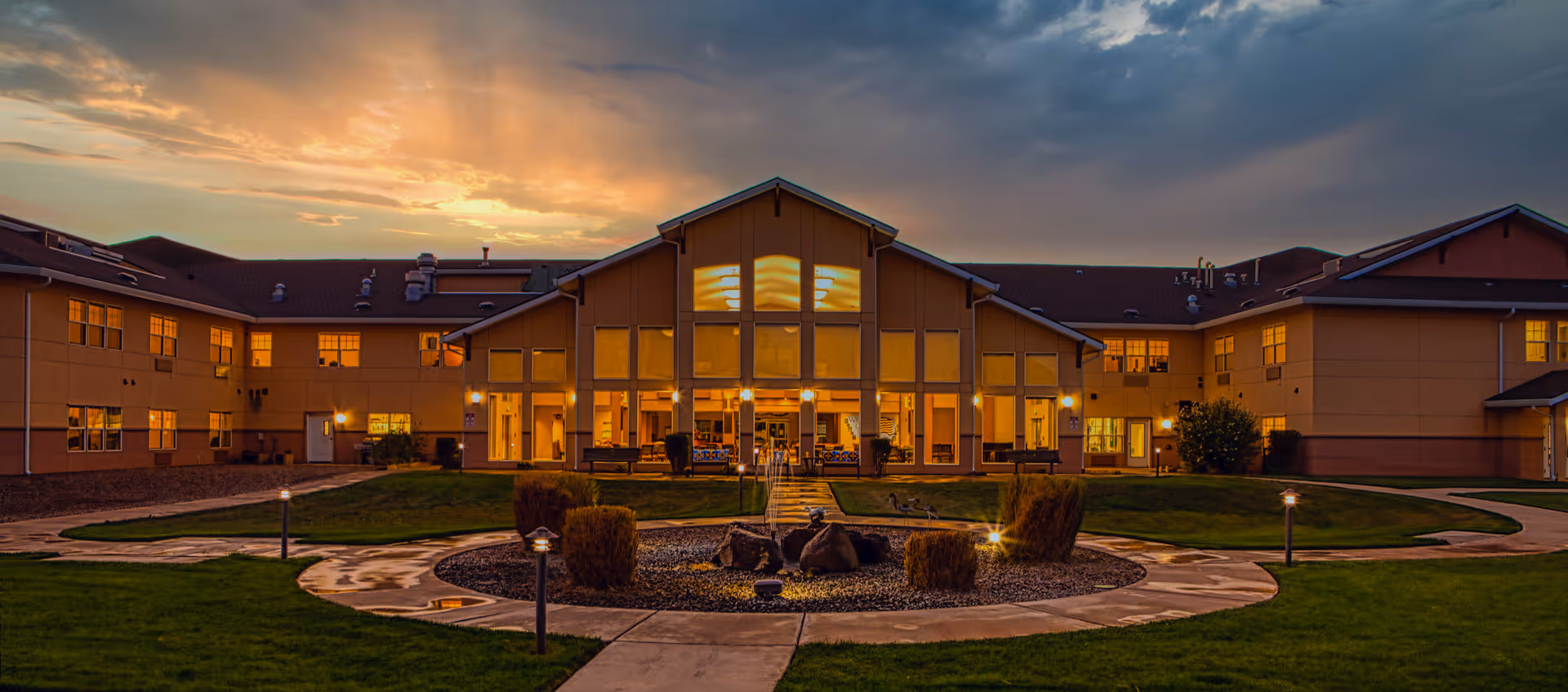 Exterior view of Wheatfields Estates Senior Living and Memory Care building at dusk, with warm lights glowing from the windows and a landscaped circular pathway with bushes and rocks in the foreground under a partly cloudy sky.