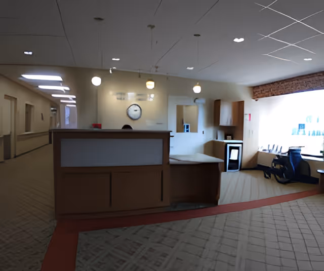 Reception area inside a senior living facility with a wooden front desk, a clock on the wall behind it, hanging pendant lights, a hallway to the left, and a seating area with chairs near a large window on the right.