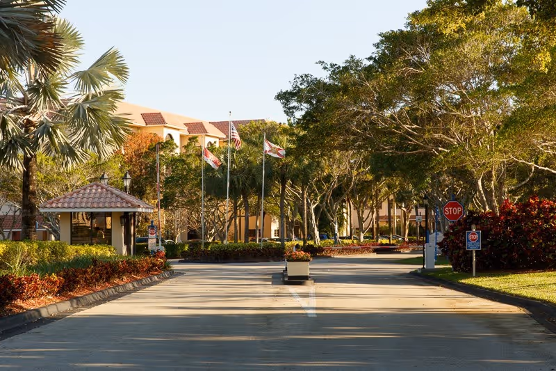 Driveway entrance to a senior living community with a guardhouse, flagpoles, trees and a stop sign.