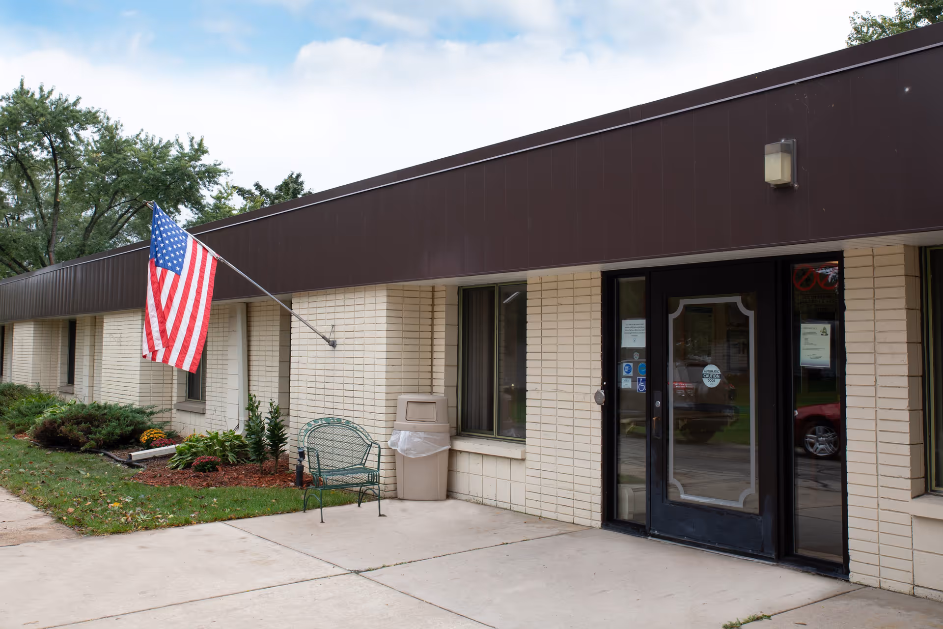 Exterior view of a single-story brick building with a dark brown roof trim. An American flag is mounted on the wall near the entrance, which has glass double doors. There is a green metal bench and a trash can near the entrance, with some landscaping including bushes and flowers along the building. Trees and a partly cloudy sky are visible in the background.