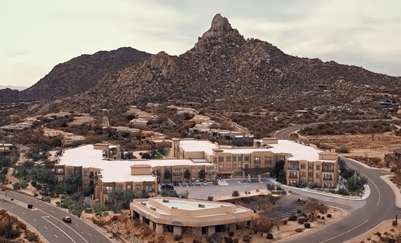 Aerial view of ACOYA Troon senior living facility surrounded by desert landscape and rocky hills, with a winding road leading to the building.