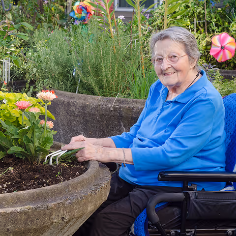 An elderly woman in a blue shirt sitting in a wheelchair and gardening outdoors, tending to flowers in a large stone planter with greenery and colorful pinwheels in the background.