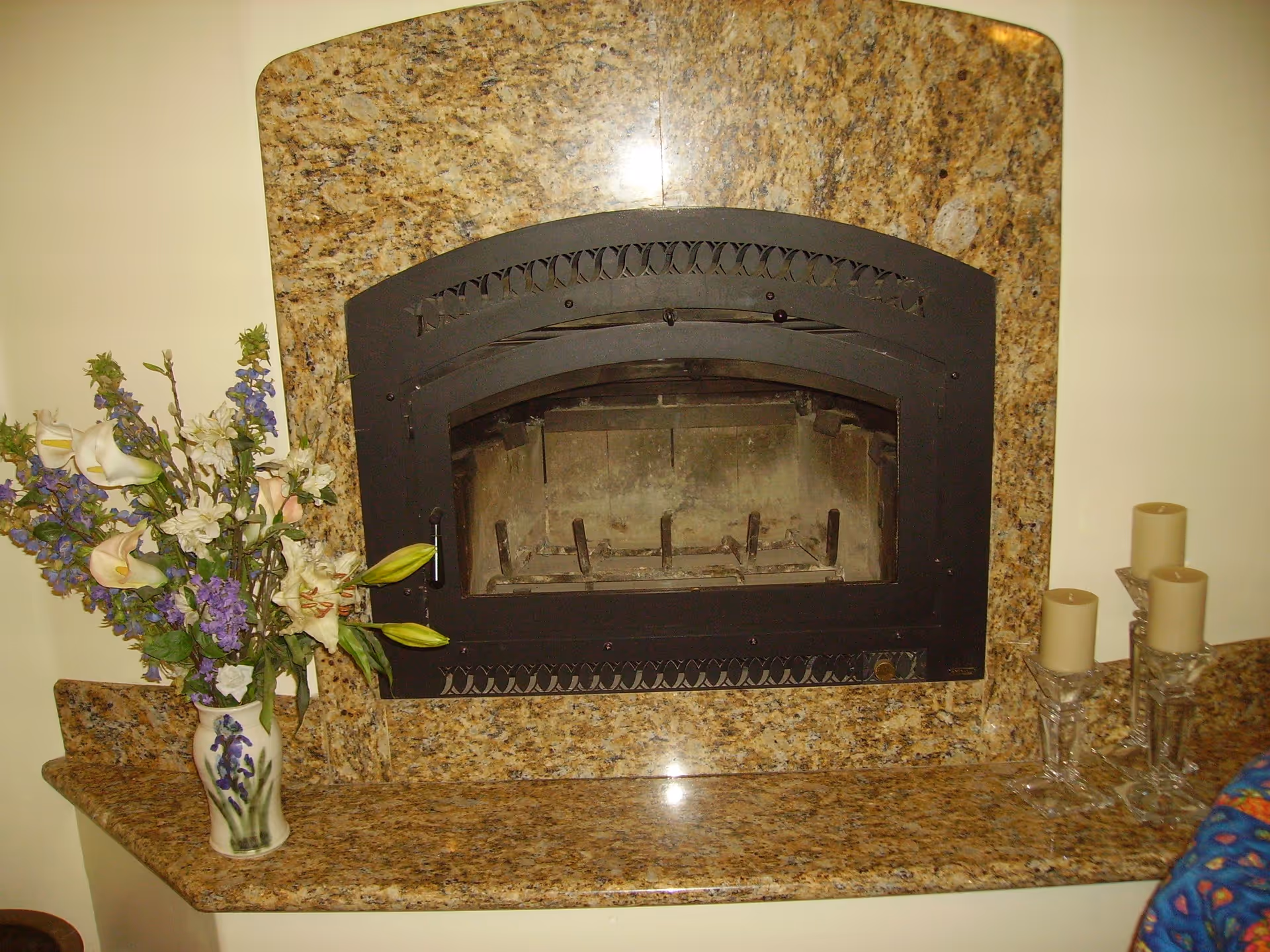 A close-up view of a stone fireplace with a black metal frame and glass door. To the left of the fireplace is a vase with a floral arrangement of white, purple, and green flowers. To the right, there are three beige candles placed on glass candle holders of varying heights.