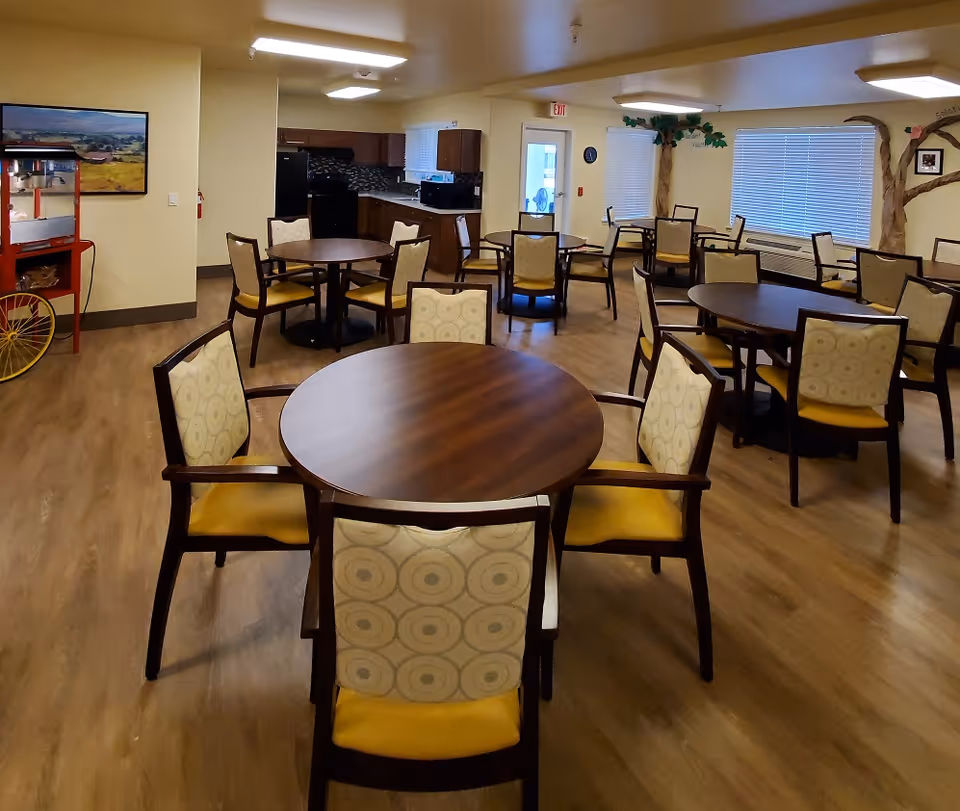 Interior dining room with round wooden tables and upholstered chairs and a kitchenette in the background.