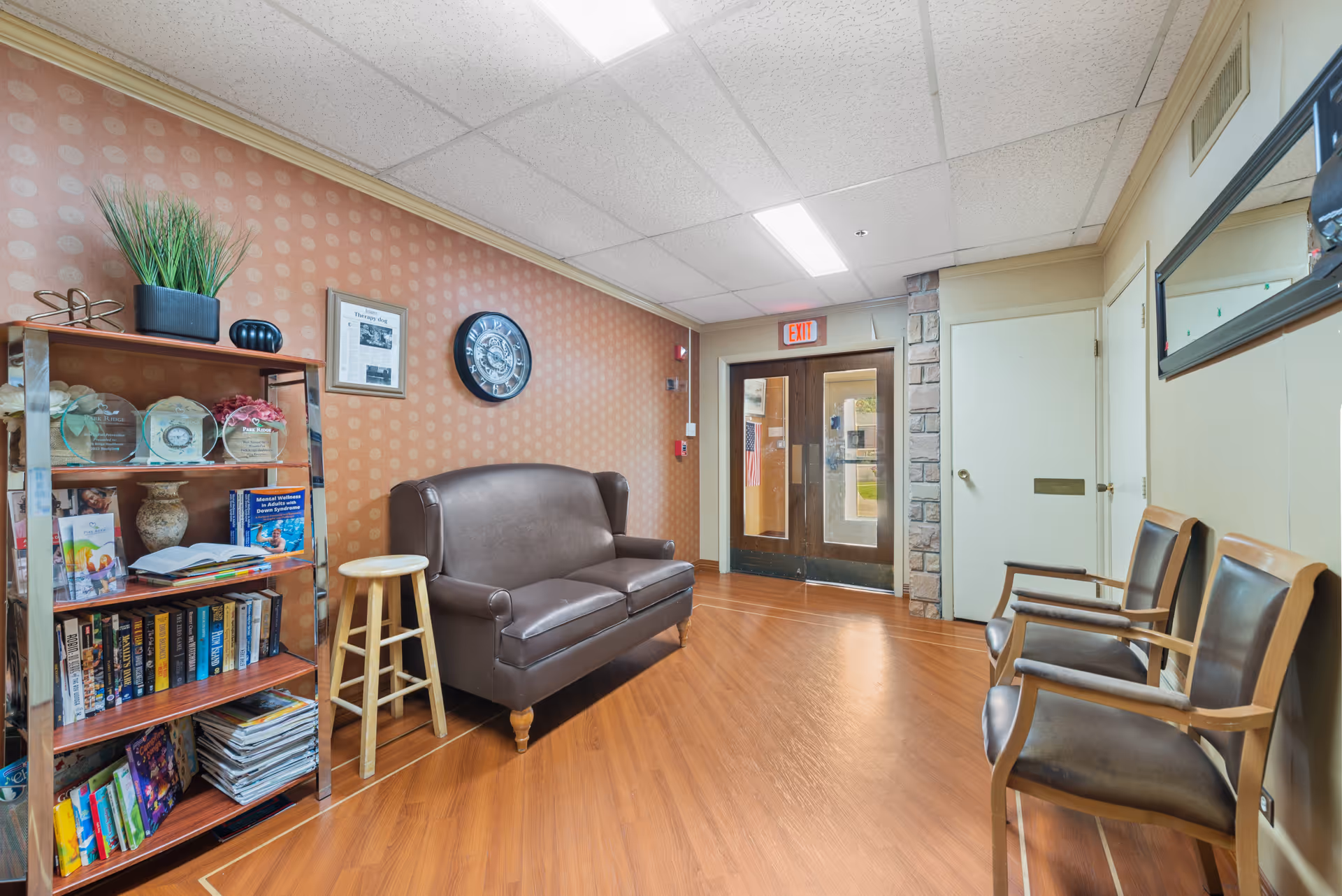 A senior living facility waiting area with a leather sofa, wooden chairs, a bookshelf, and double glass entry doors under an EXIT sign.